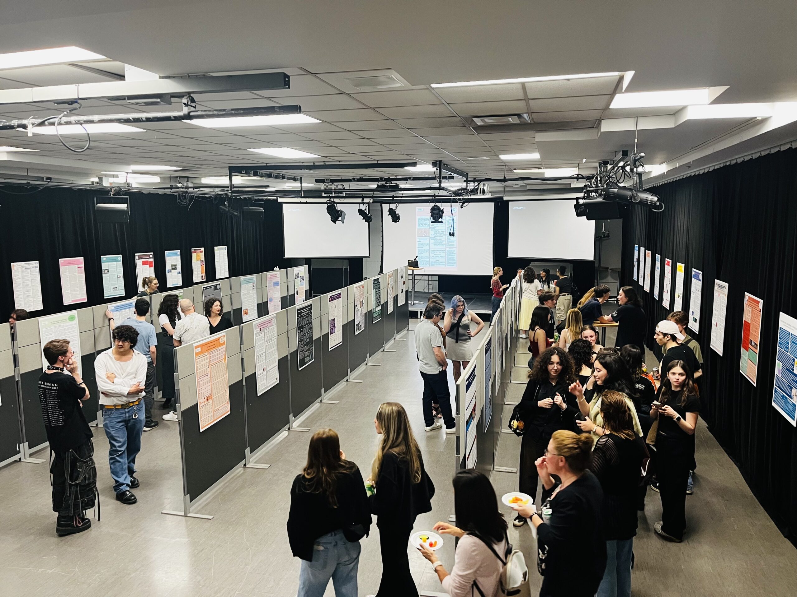 Une photo prise du haut d'une mezzanine. On y voit la salle du cégep aménagée en grande exposition d'affiches accrochées sur des longs panneaux alignés. La foule d'étudiants, professeurs et invités circulent entre les panneaux et lit les affiches en mangeant quelques bouchées.