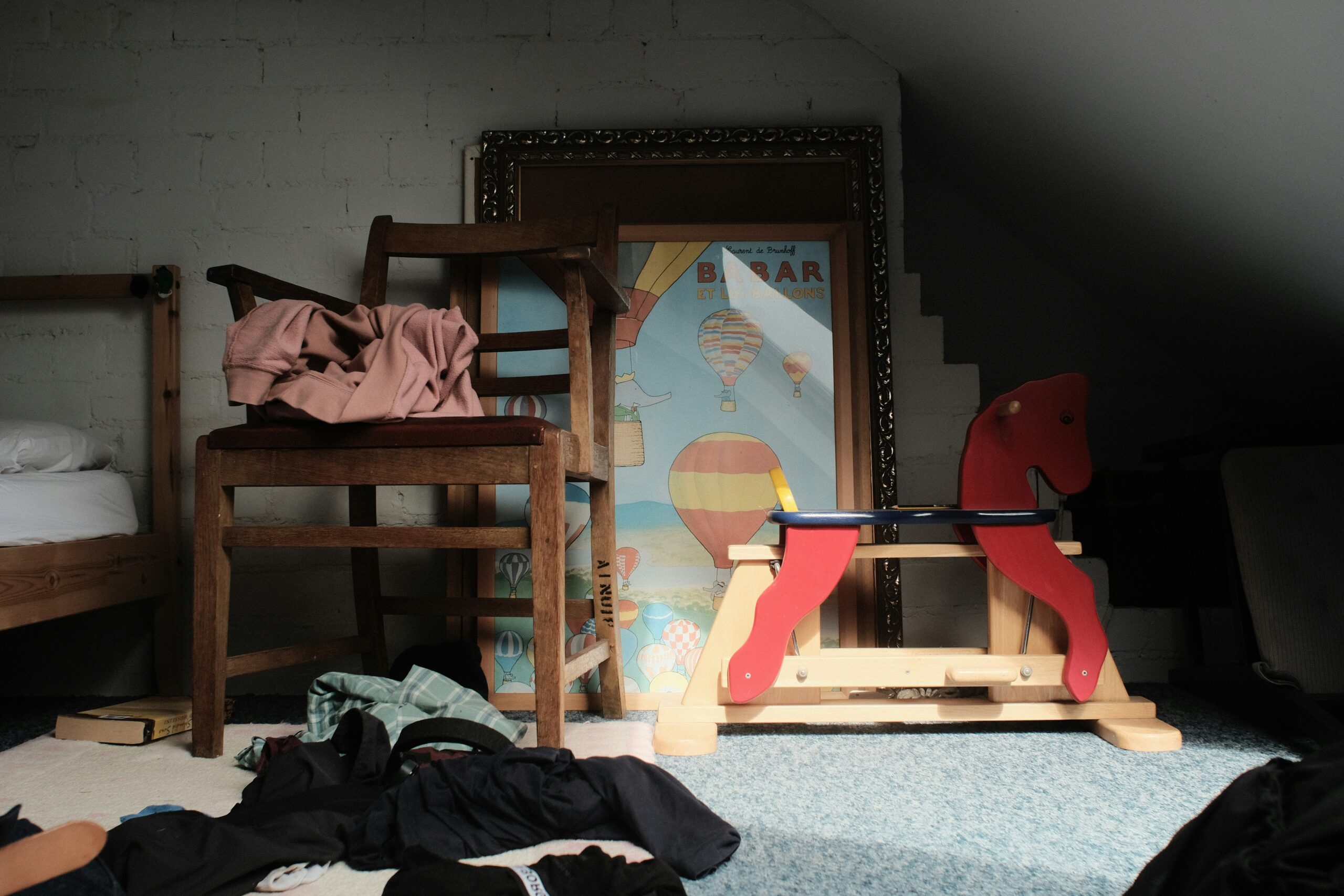 Image de couverture illustrant le rapport sur la violence domestique et le développement des enfants. La photo est prise à hauteur d'enfant ; on y voit une chaise en bois sur laquelle est posé un linge froissé. D'autres vêtements sont laissés au sol en désordre, sur la moquette de la chambre. À côté de la chaise se trouve une assise pour enfant en forme de cheval de bois. Derrière, deux cadres représentant des illustrations pour enfant sont posés au sol. La pièce est dans la pénombre et l’ambiance de la photo donne une impression de désordre silencieux.