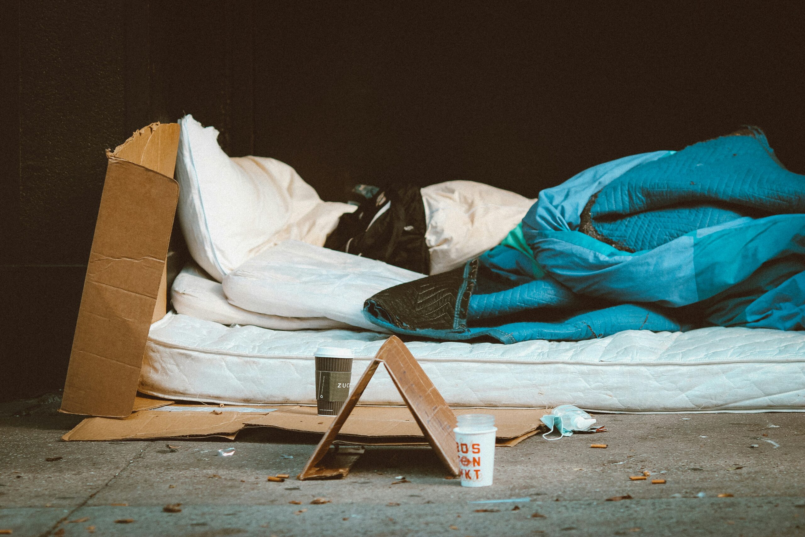 Photographie d'un campement dans la rue. Un matelas posé sur des cartons est recouvert d'un oreiller et d'une couverture. Plusieurs cup de café sont posées au sol.
